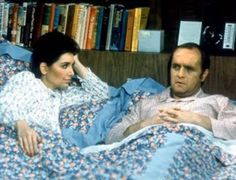 a man and woman laying in bed next to each other with books on the shelves behind them