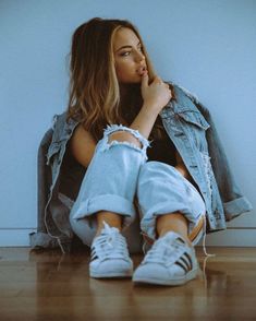 a woman sitting on the floor with her legs crossed, wearing white sneakers and denim jacket