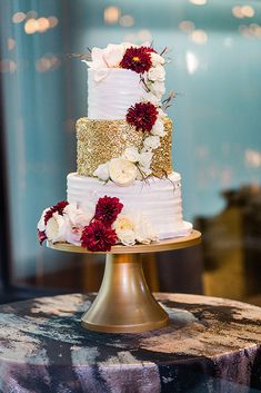 a three tiered wedding cake with red and white flowers on the top is sitting on a gold pedestal