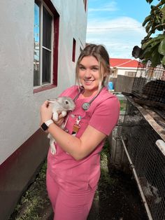 a woman in pink scrubs holding a small animal and posing for the camera with her hands on her chest