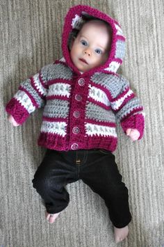 a baby wearing a pink and gray knitted jacket laying on the floor next to a pair of shoes