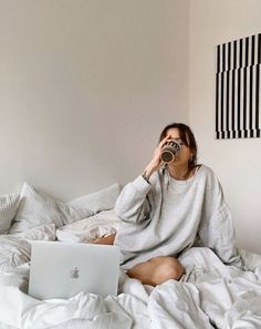 a woman sitting on her bed drinking from a cup while looking at her laptop computer