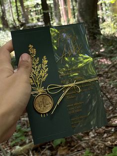 a person holding up a green and gold wedding card in the middle of some woods