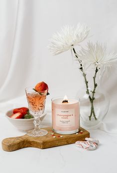 a candle and bowl of strawberries on a wooden board with flowers in the background