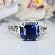 a blue and white diamond ring sitting on top of a table with flowers in the background