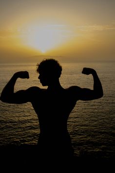 a man flexing his muscles in front of the ocean at sunset or sunrise time