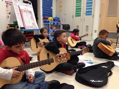 several children are sitting on the floor playing guitars in a room with other children around them