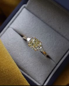 an engagement ring in a gift box on a yellow velvet surface, with the diamond center surrounded by two smaller diamonds