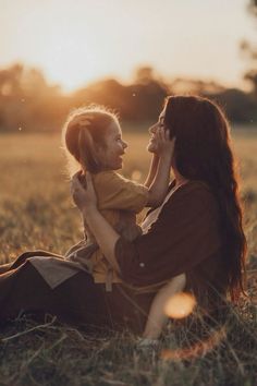 a woman holding a small child in her arms while sitting on the grass at sunset