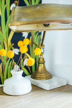 small yellow flowers in a white vase on a wooden table next to a gold lamp