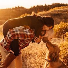 a woman kissing her dog in the middle of a field with trees and bushes behind her