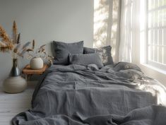 a bed with grey linens and two vases on the side table in front of it