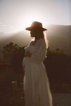 a woman in a white dress and hat looking at the sun set over mountains with trees