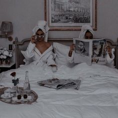 two women in white robes sitting on a bed while reading magazines and drinking tea together