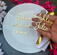 a person holding a white plate with gold nameplates on it and pink flowers in the background