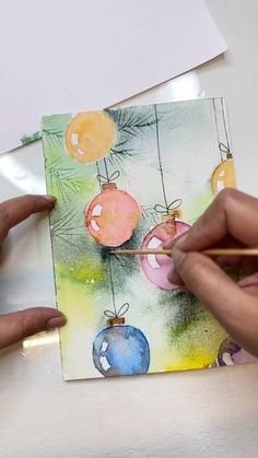 two hands are painting christmas ornaments on a piece of paper with watercolor pencils