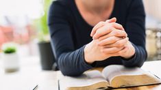 a woman sitting at a table with an open book in front of her and praying