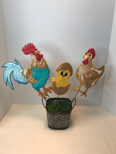 three metal roosters are standing on top of a potted plant in front of a white wall