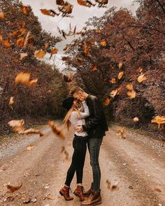 two people standing in the middle of a dirt road surrounded by leaves and falling from trees