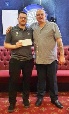 two men standing next to each other in front of a red couch with darts on it