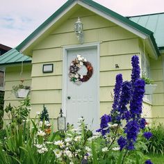 a yellow house with purple and white flowers in the foreground, and a wreath on the front door