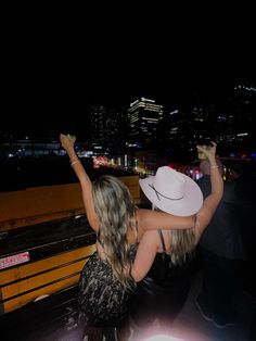 two people sitting on a bench at night with their arms in the air and one person wearing a white hat