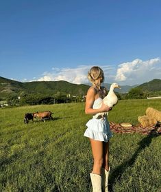 a woman is holding a goose in a field with other animals and cows behind her