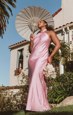 a woman in a long pink dress holding an open parasol and standing next to a house