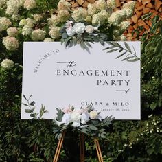 a white sign with flowers on it in front of some trees and bushes that says the engagement party