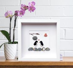 a white frame with rocks and shells in it next to a purple flower on a table