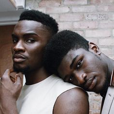 two men are posing for the camera in front of a brick wall, one is holding his arm around the other man's shoulder