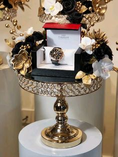 a gold and black flower arrangement on top of a white pedestal with a watch in it