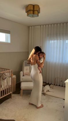 a woman in a white dress is holding a baby and looking into the mirror while standing next to a crib