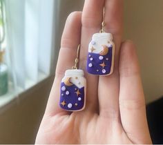 a hand holding two small earrings in it's left hand, one is purple and the other is white