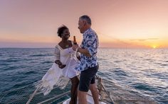 a man and woman standing on the back of a boat in the ocean at sunset