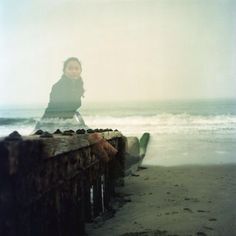 a woman sitting on top of a wooden fence next to the ocean with waves coming in