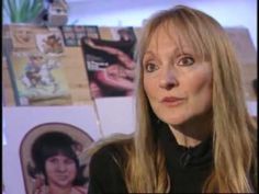 a woman with long blonde hair standing in front of pictures on the wall and looking at the camera