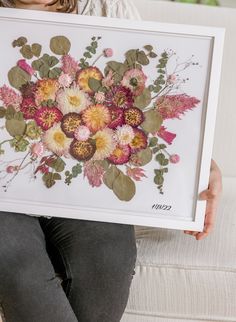 a woman sitting on a couch holding up a framed painting with flowers in the middle