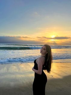 a woman in a black dress standing on the beach looking up at the sun setting