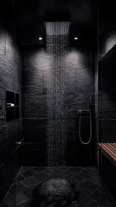 a shower head in the middle of a black tiled bathroom with wooden bench under it