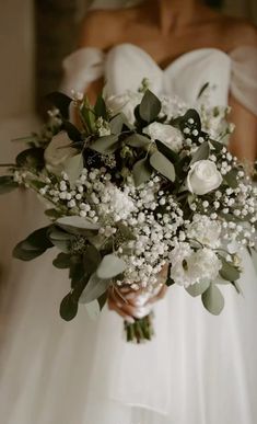 a bride holding a bouquet of white flowers and greenery in her wedding day dress