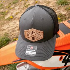 This customized hat is great for every dirt bike riding or motocross lover. The leather patch in front will come with your state letters on either side of the cross arrows and state map on the bottom. The top emblem will be the dirt bike or you can customize this by selecting another one from out options list. If you are looking for a different emblem please let us know and we will see if we can find it. This is a one size fits most, adjustable snap back Richardson original 112 trucker hat in gr Biker Cap For Outdoor Use, Biker Cap For Outdoor, Outdoor Biker Cap, Biker Style Cap For Outdoor, Adjustable Cap For Biker Events, Adjustable Black Motorcycle Hat, Custom Outdoor Baseball Cap With Flat Bill, Dirt Bike Riding, Motocross Mom