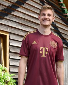 a man standing in front of a wooden building wearing a maroon and gold soccer jersey