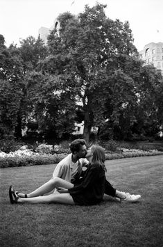 two people are sitting on the grass in front of some trees and bushes, one is kissing the other's cheek