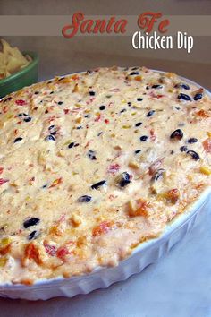 a chicken dip in a white dish on a table next to a bowl of chips