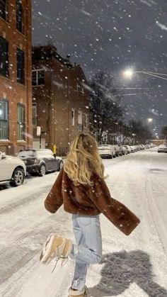 a woman is walking her dog in the snow at night, with cars parked on the street behind her