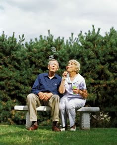 an older man and woman sitting on a bench in front of some trees blowing bubbles