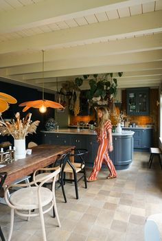 a woman walking through a kitchen next to a dining room table and two lamps hanging from the ceiling