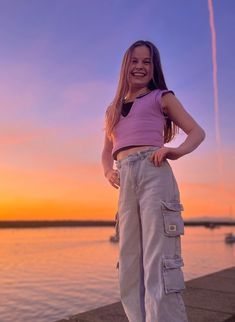 a girl standing on the edge of a pier at sunset with her hands in her pockets