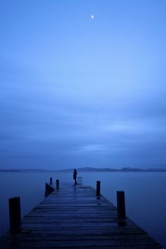 a person standing on a dock in the middle of water at night with a full moon behind them